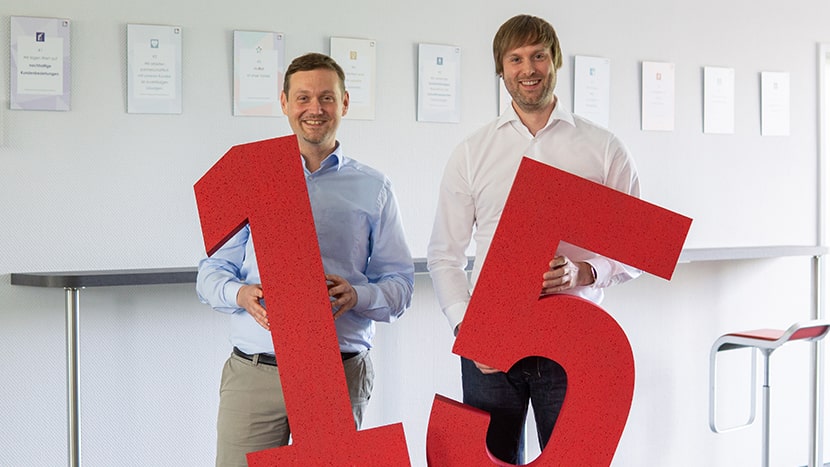 Matthias Besenfelder und Guido Nippe, die Geschäftsleitung der BROCKHAUS AG, präsentiert sich mit einer großen roten 15 zum Firmenjubiläum.